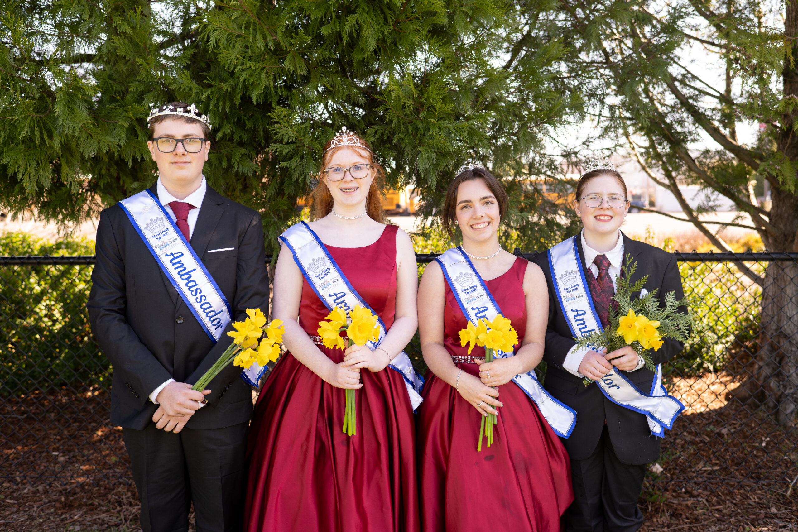 Four Fair ambassadors wearing crowns and sashes pose together with yellow flower bouquets.
