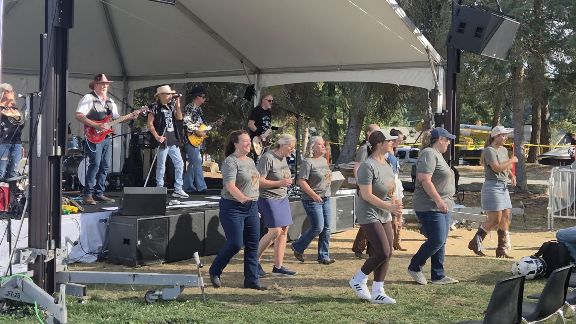 Fairgoers line dance in front of a live band on an outdoor stage.